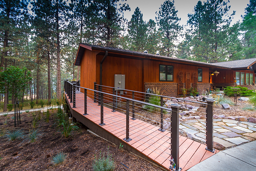 A long, expensive-looking red wood ramp curves up around the back of the house past garden features and paving stones.  The ramp has cables that prevent children and wheelchairs from falling off the ramp underneath the black metal guardrails.